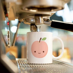 Könnte beinhalten: Weiße Keramik-Kaffeetasse mit einer niedlichen Pfirsich-Illustration, einem lächelnden Gesicht, einem grünen Blatt und einem braunen Stiel. Die Tasse steht auf einer Metalloberfläche, mit einer Kaffeemaschine im Hintergrund.