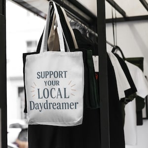 May include: A white tote bag with the text "SUPPORT YOUR LOCAL Daydreamer" in blue and light orange. The bag is hanging on a black metal rack with other clothing items. The background is blurred, suggesting a store setting.