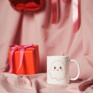 Könnte beinhalten: Weiße Keramiktasse mit einer Illustration eines lächelnden Geistes. Auf rosa Stoff, mit einem roten herzförmigen Ballon und einer roten Geschenkbox mit rosa Schleife. Valentinstag-Thema.