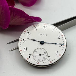 May include: A vintage Elgin watch face with a white dial, black numerals, and blue hands. The watch face has a small seconds dial and the word "ELGIN" printed on the face. The watch face is held by tweezers.