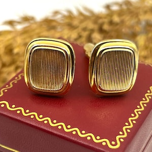 May include: A pair of gold-toned cufflinks with a rectangular, ridged design. The cufflinks are displayed on a red box with gold trim. The background features a blurred, neutral-toned element.