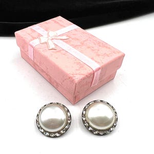 May include: Two pearl-effect stud earrings with a silver-tone border and clear crystals. A pink gift box with a ribbon and bow is in the background. The box is closed and has a textured surface.