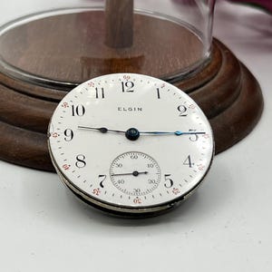 May include: An antique Elgin pocket watch face with a white dial, black numerals, and blue hands. The watch face includes a smaller subdial and the word "ELGIN". The watch face is set against a wooden base.