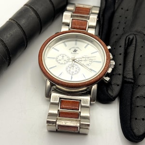May include: A silver wristwatch with a white face and brown wood accents. The watch features three sub-dials and the text "Beverly Hills Polo Club". A black golf glove and a portion of a golf club are also visible.