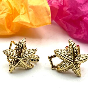 May include: A pair of gold-colored starfish-shaped earrings. The earrings have a textured surface and a post-and-clasp closure. The background features orange and pink tissue paper.