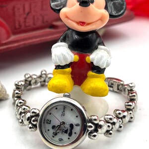 Op de afbeelding: Een zilverkleurig Disney-horloge met een witte wijzerplaat met Mickey Mouse. De horlogeband heeft een uniek kralenontwerp. Een Mickey Mouse-beeldje in klassieke kleuren zit boven het horloge. Een rode speelgoedbrandweerwagen staat op de achtergrond.