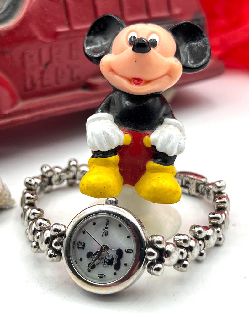 Op de afbeelding: Een zilverkleurig Disney-horloge met een witte wijzerplaat met Mickey Mouse. De horlogeband heeft een uniek kralenontwerp. Een Mickey Mouse-beeldje in klassieke kleuren zit boven het horloge. Een rode speelgoedbrandweerwagen staat op de achtergrond.