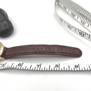 May include: A brown leather watch band with a gold-colored watch face. The band is measured with a white measuring tape. The watch band is approximately 3 inches long.