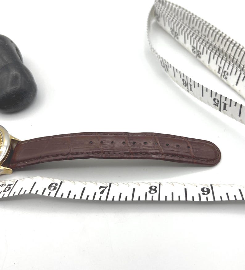 May include: A brown leather watch band with a gold-colored watch face. The band is measured with a white measuring tape. The watch band is approximately 3 inches long.