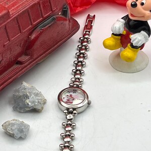 Op de afbeelding: Een zilverkleurig horloge met een Mickey Mouse-wijzerplaat en een schakelband. Het horloge staat naast een rode speelgoedbrandweerwagen en een Mickey Mouse-beeldje. Twee lichtblauwe kristallen zijn ook zichtbaar.