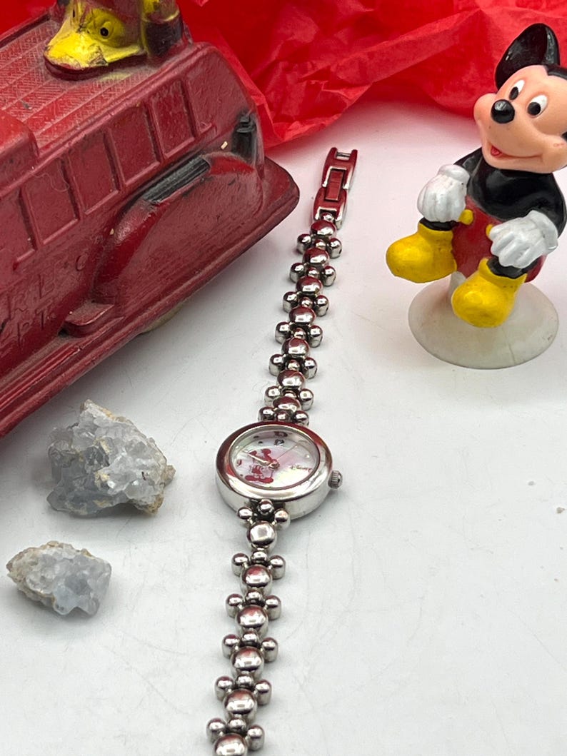 Op de afbeelding: Een zilverkleurig horloge met een Mickey Mouse-wijzerplaat en een schakelband. Het horloge staat naast een rode speelgoedbrandweerwagen en een Mickey Mouse-beeldje. Twee lichtblauwe kristallen zijn ook zichtbaar.