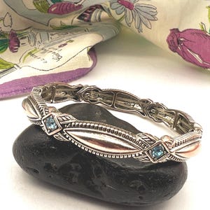 Può includere: Bracciale argentato dal design ornato, con forme a foglia e piccole gemme blu quadrate. Il bracciale è appoggiato su una pietra scura e strutturata, con un tessuto a motivi floreali sullo sfondo.