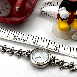 Op de afbeelding: Zilverkleurig Disney-horloge met een parelmoeren wijzerplaat en een Mickey Mouse-ontwerp. De horlogeband heeft een uniek, bubbelachtig ontwerp. Een witte meetlint ligt naast het horloge. De diameter van het horloge is ongeveer 2,5 cm.