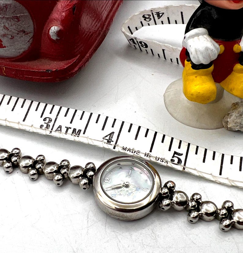 Op de afbeelding: Zilverkleurig Disney-horloge met een parelmoeren wijzerplaat en een Mickey Mouse-ontwerp. De horlogeband heeft een uniek, bubbelachtig ontwerp. Een witte meetlint ligt naast het horloge. De diameter van het horloge is ongeveer 2,5 cm.