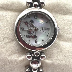 Op de afbeelding: Zilverkleurig Disney-horloge met een ronde wijzerplaat en een Mickey Mouse-ontwerp. Het horloge heeft een parelmoeren wijzerplaat met zilveren wijzers en uurmarkeringen. De band is gemaakt van verbonden zilveren kralen. Het woord "Disney" staat op de wijzerplaat.