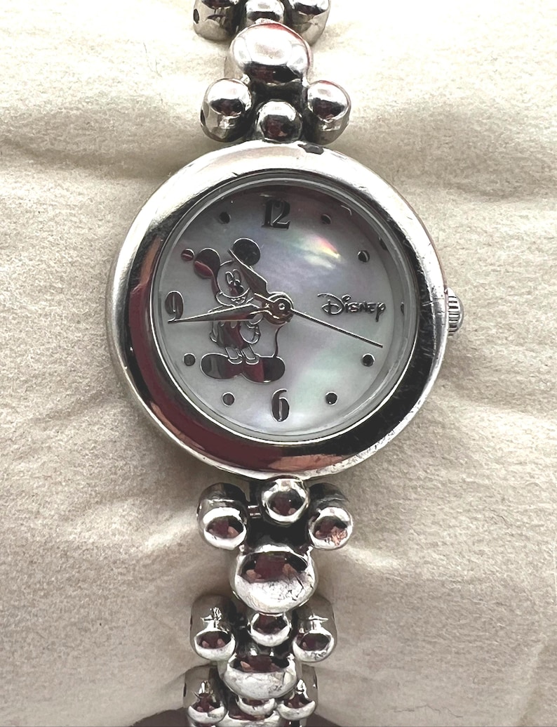 Op de afbeelding: Zilverkleurig Disney-horloge met een ronde wijzerplaat en een Mickey Mouse-ontwerp. Het horloge heeft een parelmoeren wijzerplaat met zilveren wijzers en uurmarkeringen. De band is gemaakt van verbonden zilveren kralen. Het woord "Disney" staat op de wijzerplaat.