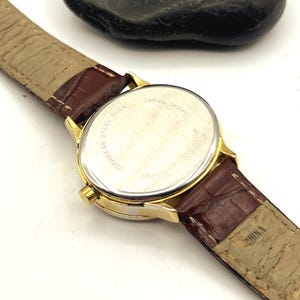 May include: A gold-toned wristwatch with a brown leather band. The watch face is visible, with the text "STAINLESS STEEL BACK" and "JAPAN MOVT". The band is brown with a light tan underside.