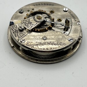 May include: Close-up of a vintage Elgin Rail Watch Co. pocket watch movement. The silver-toned mechanism features intricate gears, the text "15 Jewels", and the serial number 13418900. The watch is marked "U.S.A."