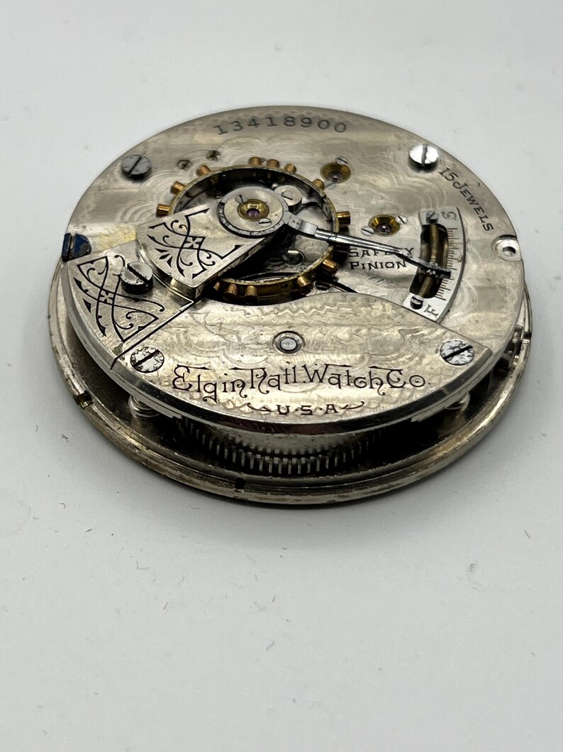 May include: Close-up of a vintage Elgin Rail Watch Co. pocket watch movement. The silver-toned mechanism features intricate gears, the text "15 Jewels", and the serial number 13418900. The watch is marked "U.S.A."