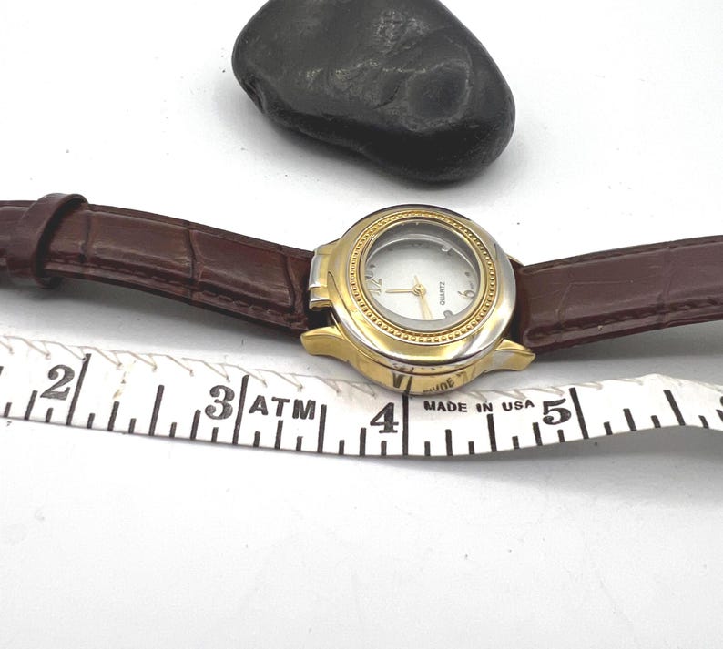 May include: A gold and silver quartz watch with a white face and brown leather band. The watch is next to a measuring tape and a dark gray stone. The watch is marked "MADE IN USA".