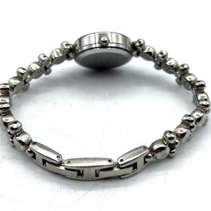 Op de afbeelding: Een zilverkleurig polshorloge met een ronde wijzerplaat en een unieke armband. De band heeft een reeks kleine, decoratieve elementen en rechthoekige schakels. Het horloge is ontworpen om om de pols te worden gedragen.