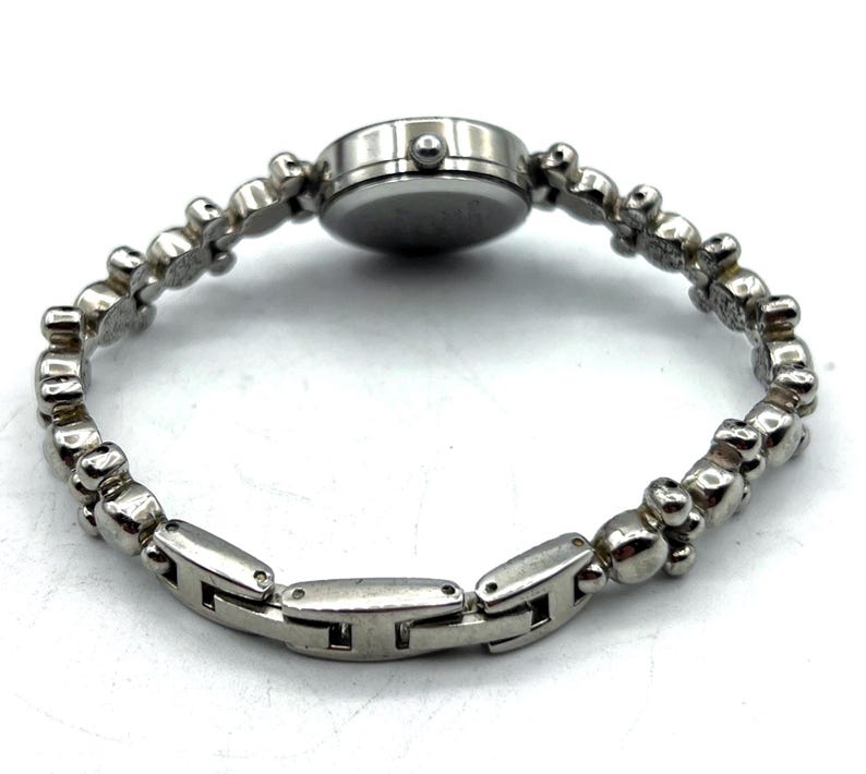 Op de afbeelding: Een zilverkleurig polshorloge met een ronde wijzerplaat en een unieke armband. De band heeft een reeks kleine, decoratieve elementen en rechthoekige schakels. Het horloge is ontworpen om om de pols te worden gedragen.