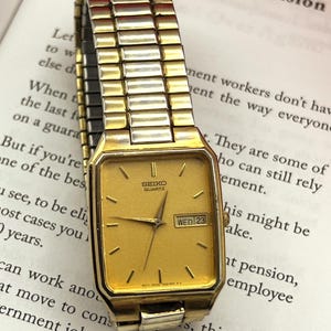 May include: A gold-tone Seiko Quartz wristwatch with a rectangular face, gold dial, and matching flexible metal expansion band. The watch features thin gold hour markers, gold hands, and a day-date window at the three o'clock position.