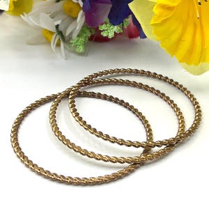 Puede incluir: Tres brazaletes dorados de metal retorcido. Los brazaletes son circulares y tienen una textura similar a una cuerda. Un ramo de flores artificiales de colores está en el fondo.