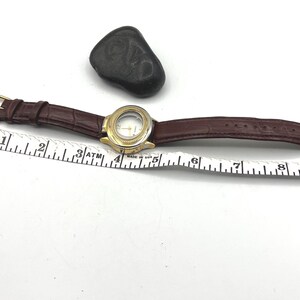 May include: A gold-tone wristwatch with a brown leather band. The watch face is clear, and the band has a crocodile texture. A black stone sits near the watch. A ruler is placed next to the watch, measuring in inches.