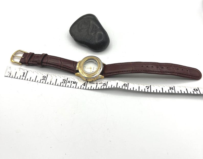 May include: A gold-tone wristwatch with a brown leather band. The watch face is clear, and the band has a crocodile texture. A black stone sits near the watch. A ruler is placed next to the watch, measuring in inches.