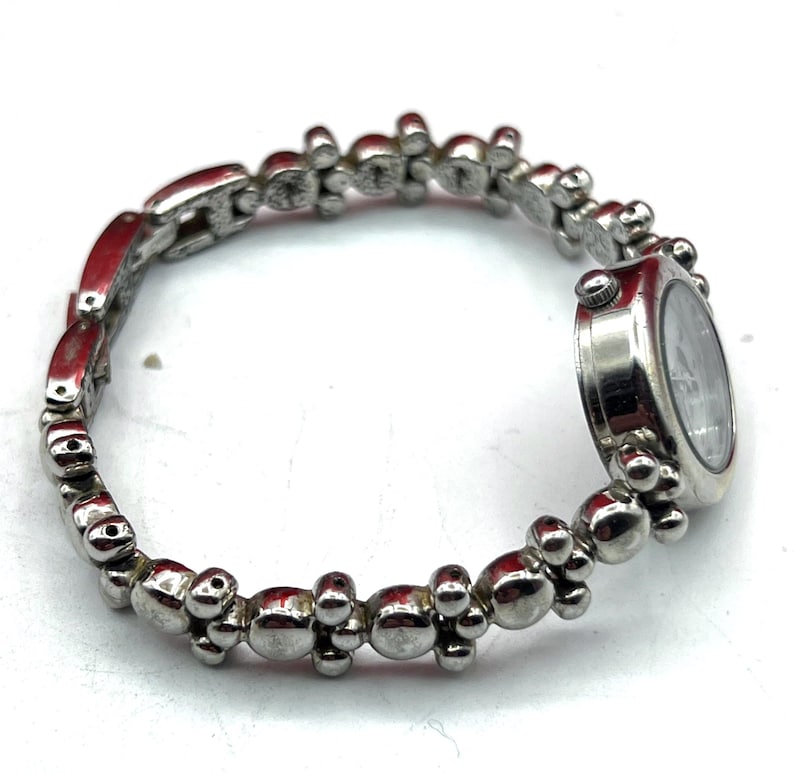 Op de afbeelding: Zilverkleurig armbandhorloge met een ronde wijzerplaat. De horlogeband heeft een reeks kleine, afgeronde en langwerpige schakels. De wijzerplaat heeft een lichte achtergrond met donkere wijzers en markeringen.
