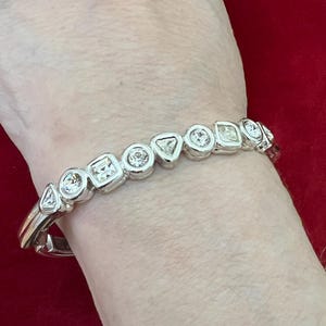 May include: A silver bracelet with a repeating pattern of geometric shapes, such as triangles, squares, and circles, each holding a clear stone. The bracelet is worn on a wrist against a red backdrop.