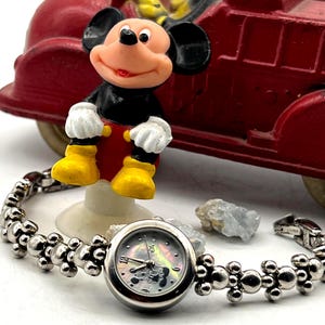 Op de afbeelding: Een zilverkleurig polshorloge met een Mickey Mouse wijzerplaat en een parelmoer achtergrond. De horlogeband heeft een uniek ontwerp met bolvormige accenten. Een Mickey Mouse beeldje en een rode speelgoedauto staan op de achtergrond.