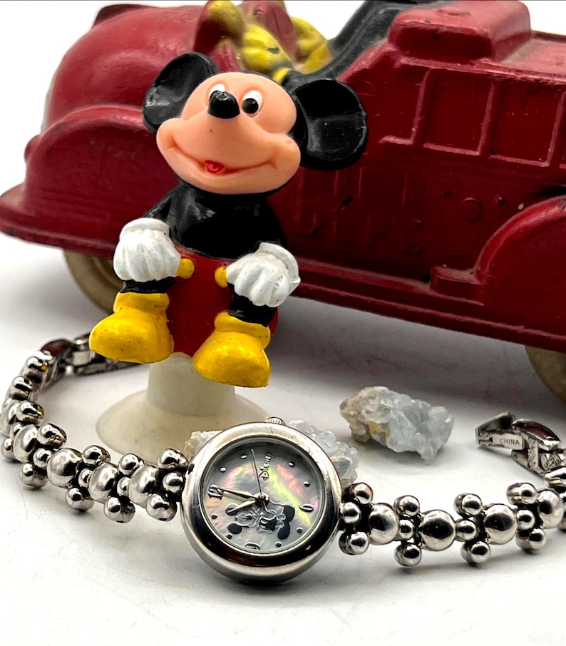 Op de afbeelding: Een zilverkleurig polshorloge met een Mickey Mouse wijzerplaat en een parelmoer achtergrond. De horlogeband heeft een uniek ontwerp met bolvormige accenten. Een Mickey Mouse beeldje en een rode speelgoedauto staan op de achtergrond.