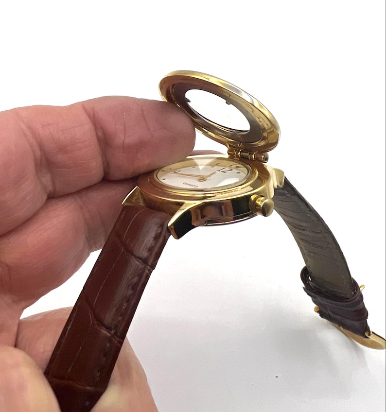 May include: A gold-toned quartz watch with a brown leather band. The watch face is white with gold hands and numerals. The watch has a hinged cover that is open, revealing the watch face. The band is a dark brown color.