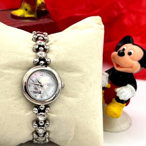 Op de afbeelding: Zilverkleurig polshorloge met een Mickey Mouse-ontwerp op de wijzerplaat en een bijpassende armband. Het horloge wordt gepresenteerd op een cr&egrave;mekleurig kussen, met een Mickey Mouse-beeldje op de achtergrond.