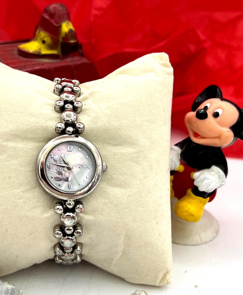 Op de afbeelding: Zilverkleurig polshorloge met een Mickey Mouse-ontwerp op de wijzerplaat en een bijpassende armband. Het horloge wordt gepresenteerd op een cr&egrave;mekleurig kussen, met een Mickey Mouse-beeldje op de achtergrond.