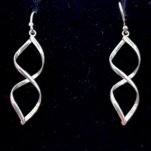 May include: A pair of silver earrings featuring a twisted infinity symbol design. The earrings hang from a small hook and are set against a black backdrop. The earrings are approximately 5 cm long.