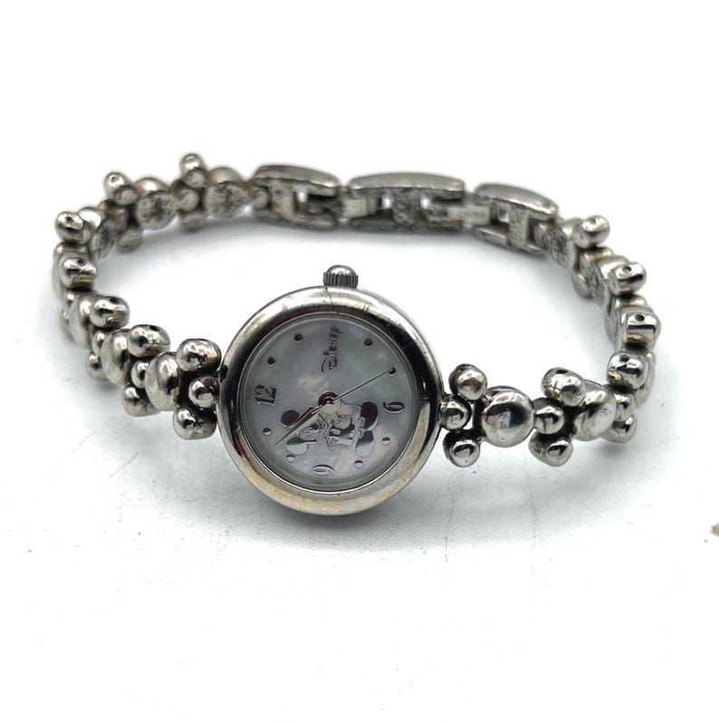Op de afbeelding: Zilverkleurig Disney-horloge met een ronde wijzerplaat met Mickey Mouse. Het horloge heeft een band in kralenarmbandstijl. De wijzerplaat heeft een parelmoer achtergrond met zilveren wijzers en cijfers. Een klassiek Disney-thema-accessoire.