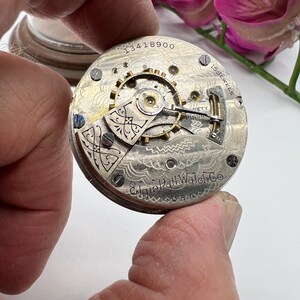 May include: Close-up of an antique Elgin Hall Watch Co. pocket watch movement. The silver-toned mechanism features intricate gears, screws, and the text "15 Jewels" and "USA". The serial number "13418900" is visible.