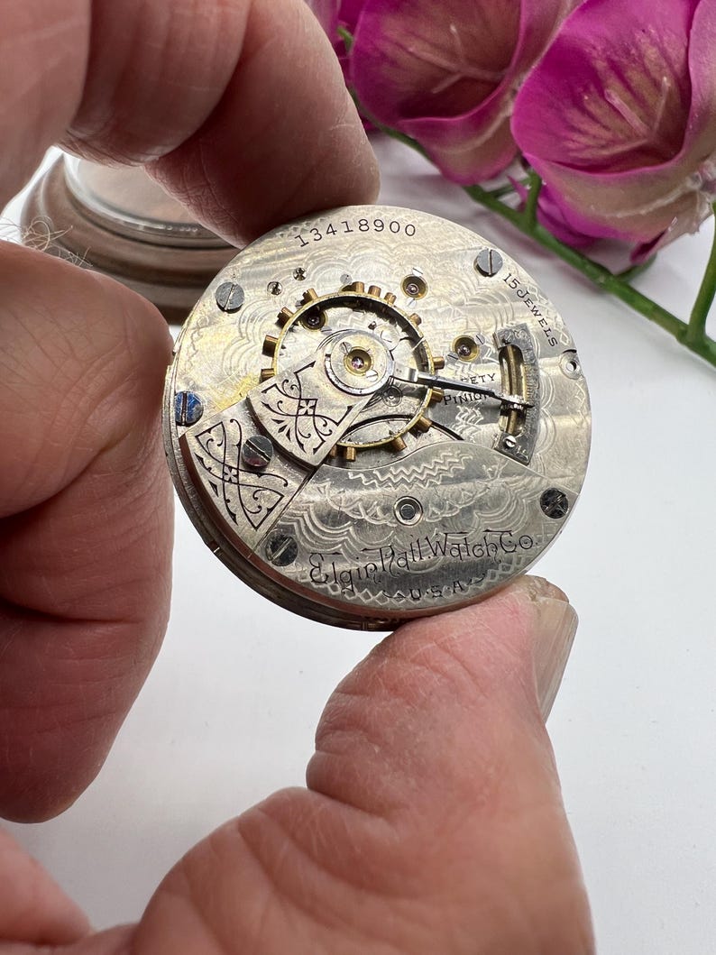 May include: Close-up of an antique Elgin Hall Watch Co. pocket watch movement. The silver-toned mechanism features intricate gears, screws, and the text "15 Jewels" and "USA". The serial number "13418900" is visible.