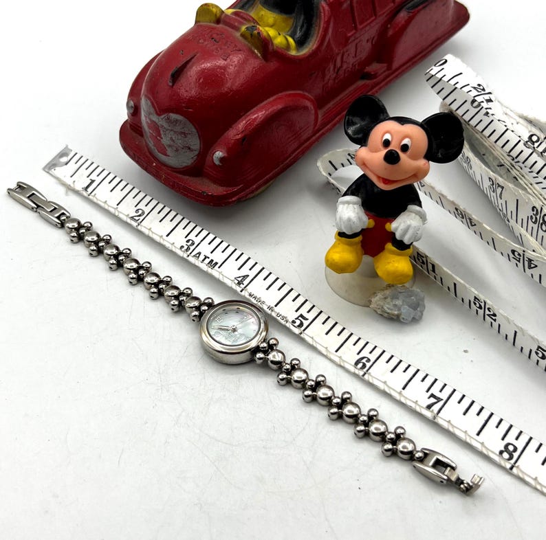 Op de afbeelding: Een zilverkleurig horloge met een ronde wijzerplaat en een kralenband wordt getoond met een Mickey Mouse-beeldje, een rode speelgoedauto en een meetlint. De wijzerplaat van het horloge heeft een lichtgekleurde, mogelijk parelmoerachtige uitstraling.