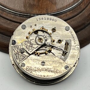 May include: Close-up of a vintage Elgin Rail Watch Co. pocket watch movement. The silver-toned mechanism features intricate gears, the text "15 Jewels", and the serial number "13418900". The watch is marked "USA".