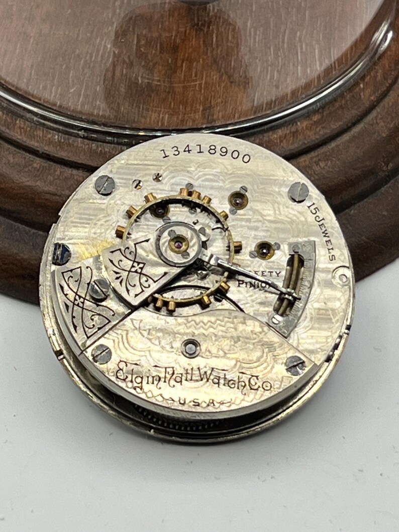 May include: Close-up of a vintage Elgin Rail Watch Co. pocket watch movement. The silver-toned mechanism features intricate gears, the text "15 Jewels", and the serial number "13418900". The watch is marked "USA".