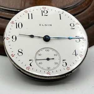 May include: A vintage Elgin pocket watch face with a white dial, black numerals, and a smaller seconds dial. The watch face has black and blue hands and the brand name "ELGIN" is printed on the dial.