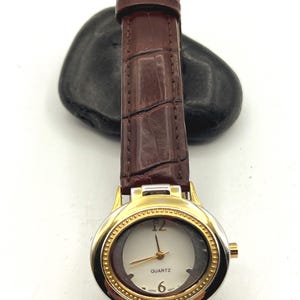 May include: A gold-toned quartz watch with a brown leather band. The watch face is white with gold hands and a gold bezel. The band is a dark brown with a crocodile texture. The watch is a classic style.