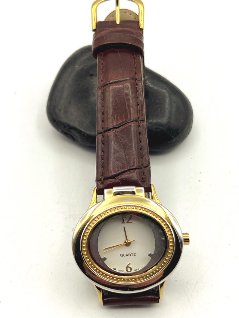 May include: A gold-toned quartz watch with a brown leather band. The watch face is white with gold hands and a gold bezel. The band is a dark brown with a crocodile texture. The watch is a classic style.