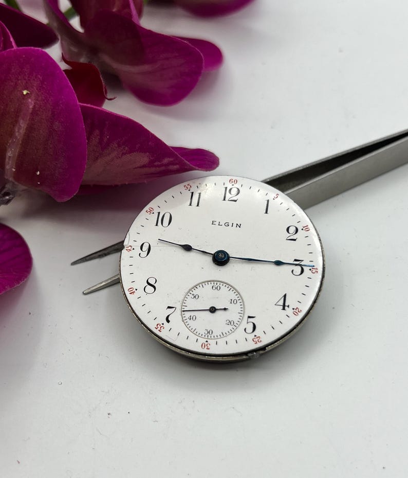 May include: A vintage Elgin watch face with a white dial, black numerals, and blue hands. The watch face has a small seconds dial and the word "ELGIN" printed on the face. The watch face is held by tweezers.