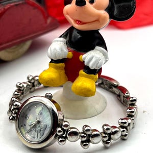 Op de afbeelding: Een zilverkleurig Mickey Mouse-horloge met een ronde wijzerplaat en een decoratieve band. De wijzerplaat heeft een parelmoer-achtige uitstraling met Mickey Mouse-afbeeldingen. Een Mickey Mouse-beeldje staat op de achtergrond, gekleed in zwart, rood, geel en wit.