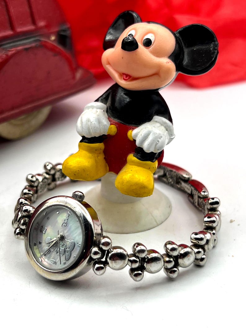 Op de afbeelding: Een zilverkleurig Mickey Mouse-horloge met een ronde wijzerplaat en een decoratieve band. De wijzerplaat heeft een parelmoer-achtige uitstraling met Mickey Mouse-afbeeldingen. Een Mickey Mouse-beeldje staat op de achtergrond, gekleed in zwart, rood, geel en wit.